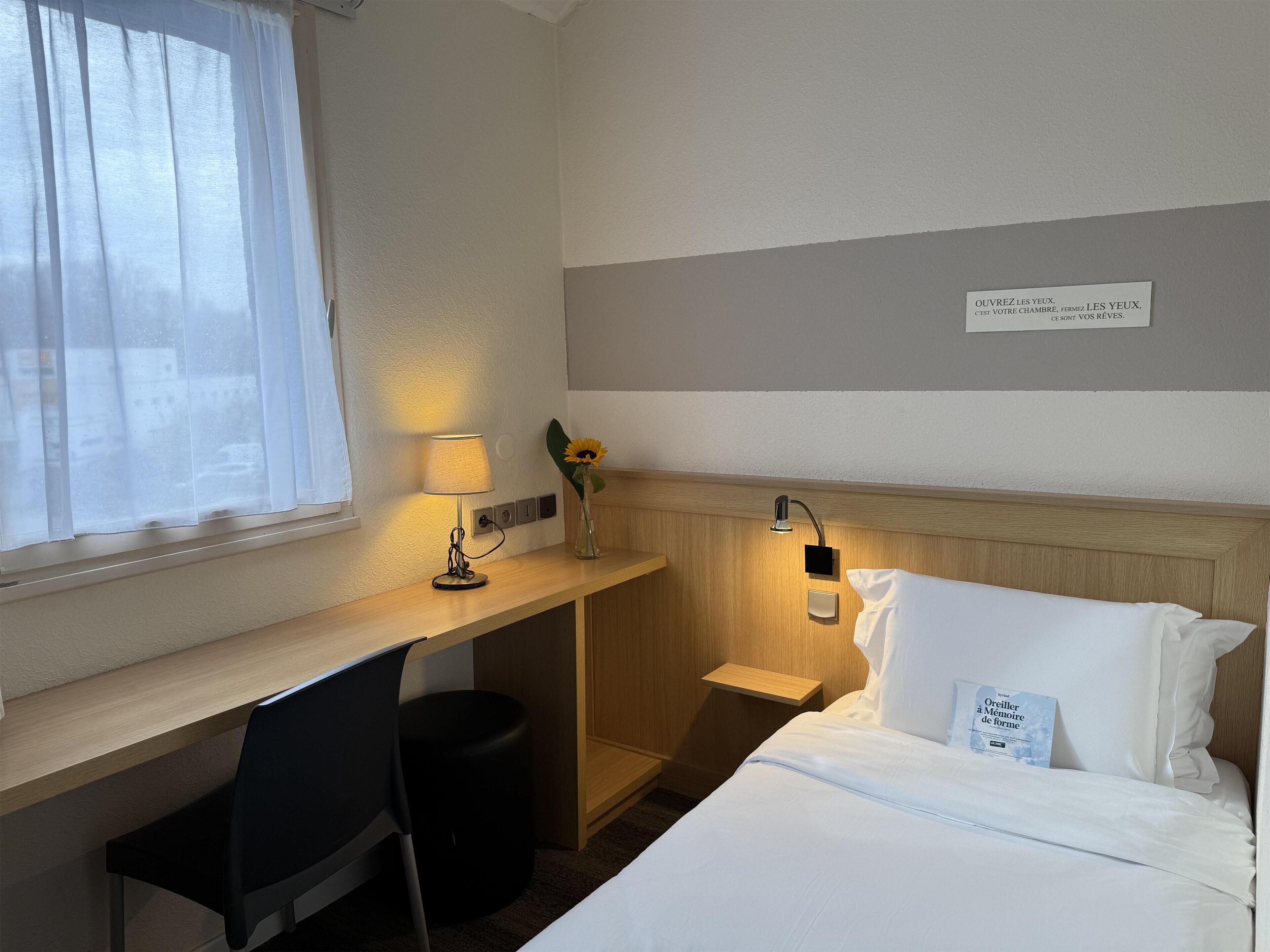 Une chambre d’hôtel comprenant un lit, un bureau avec une lampe, une chaise, une petite poubelle et une veilleuse fixée au mur.