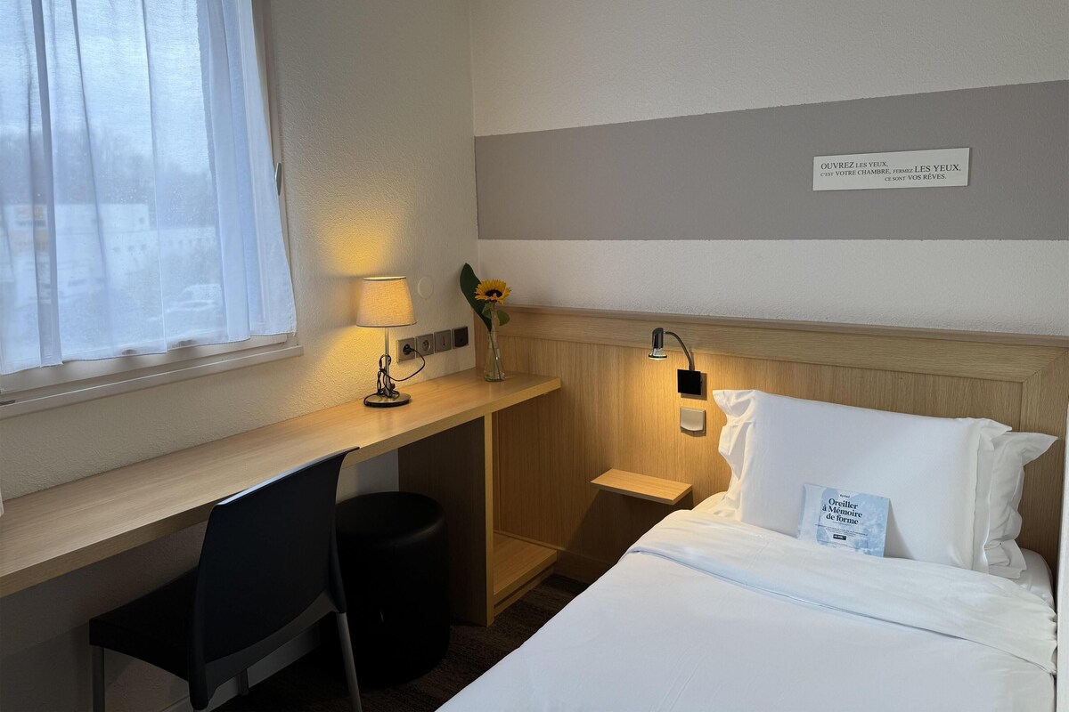 Habitación de hotel con cama, escritorio con lámpara, una silla, un cesto de basura pequeño y una luz nocturna en la pared.