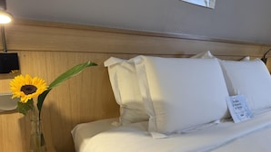 Habitación de hotel con cabecera de madera, mesita de noche con lámpara, jarrón con girasol y cama con ropa de cama y almohadas blancas.