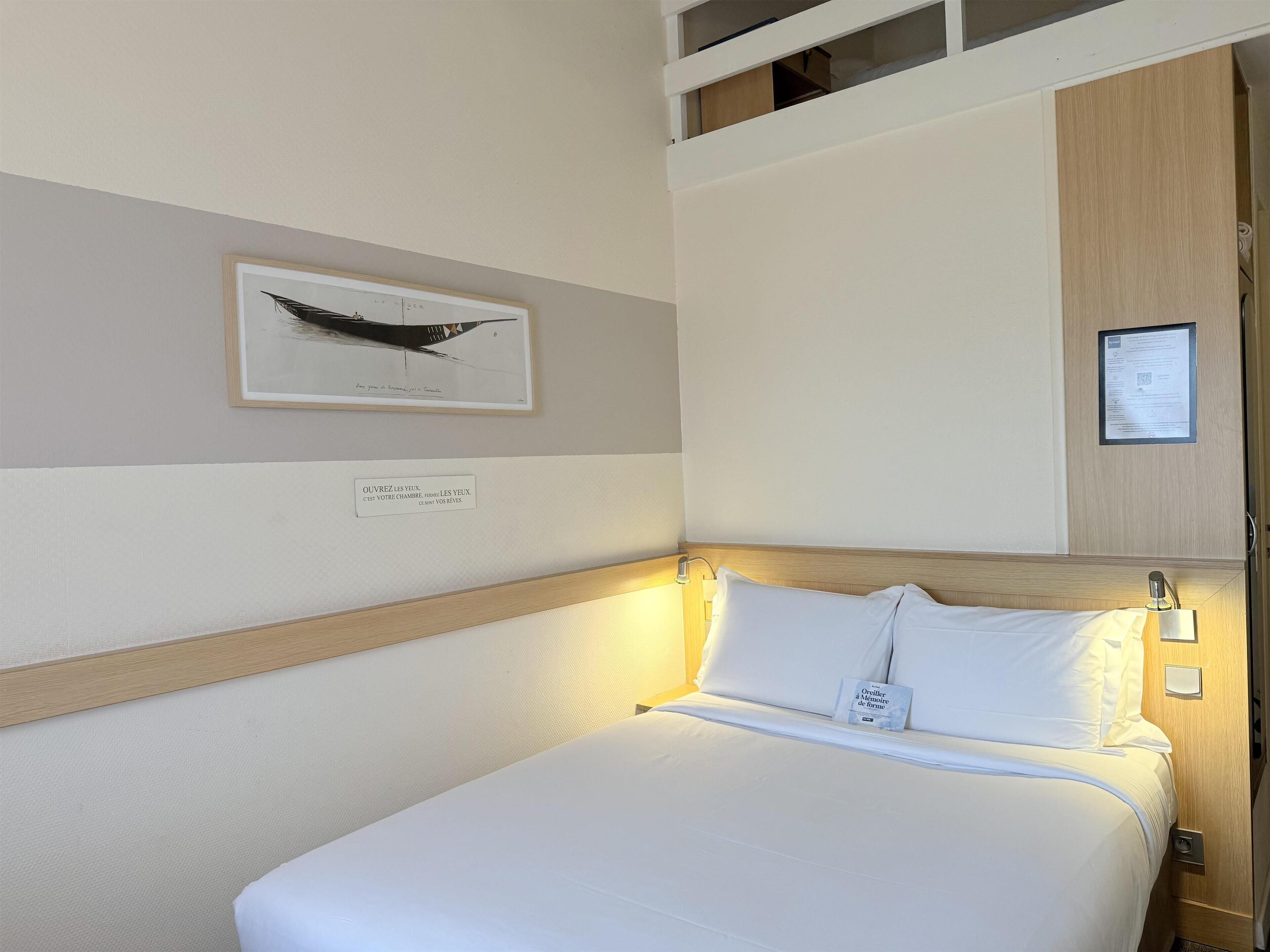 Una habitación de hotel con una cama bien hecha, una obra de arte enmarcada sobre la cama y un panel de madera en la pared.