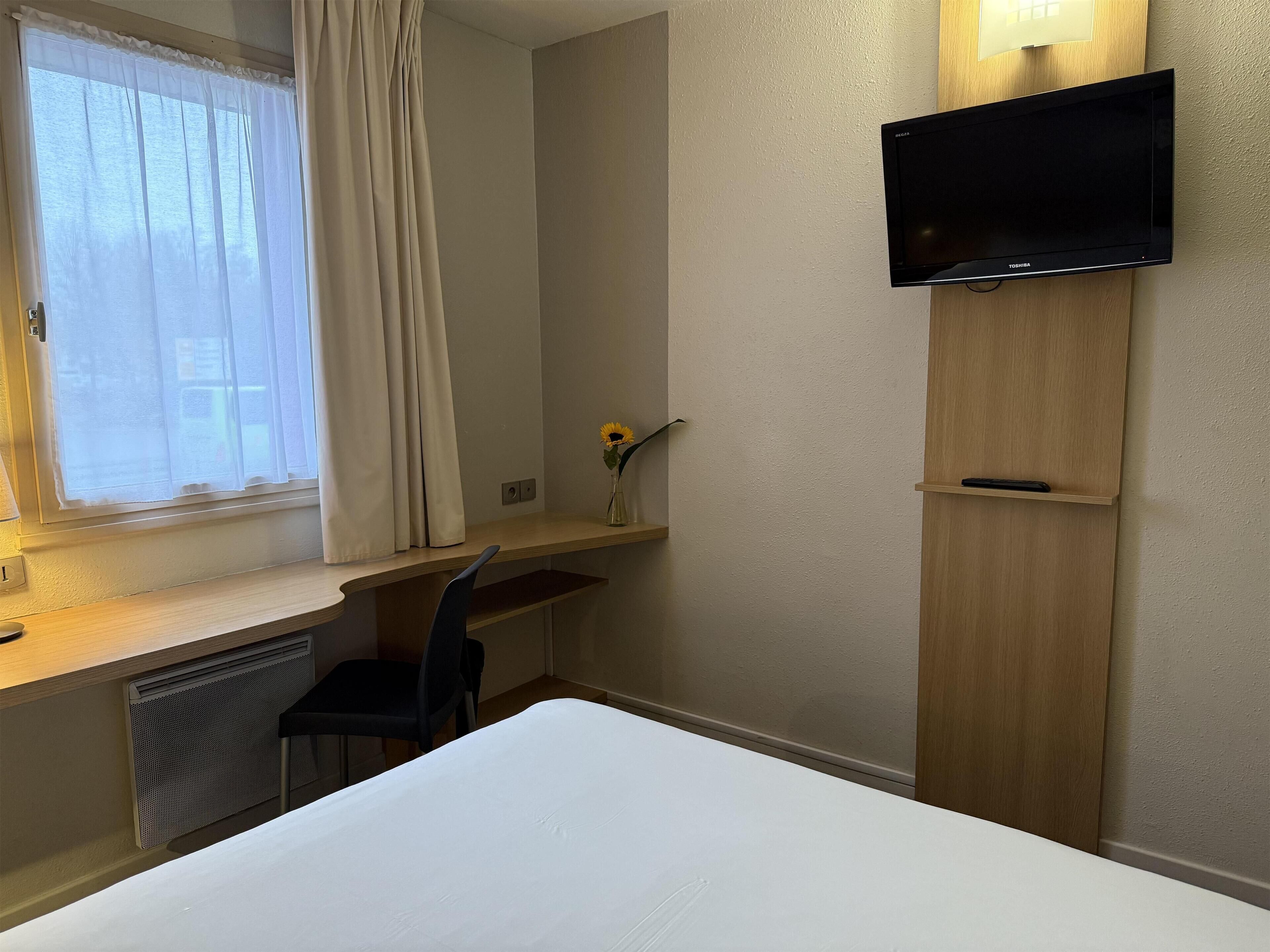 Habitación de hotel con cama, escritorio, silla, televisor montado en la pared y ventana con cortinas.