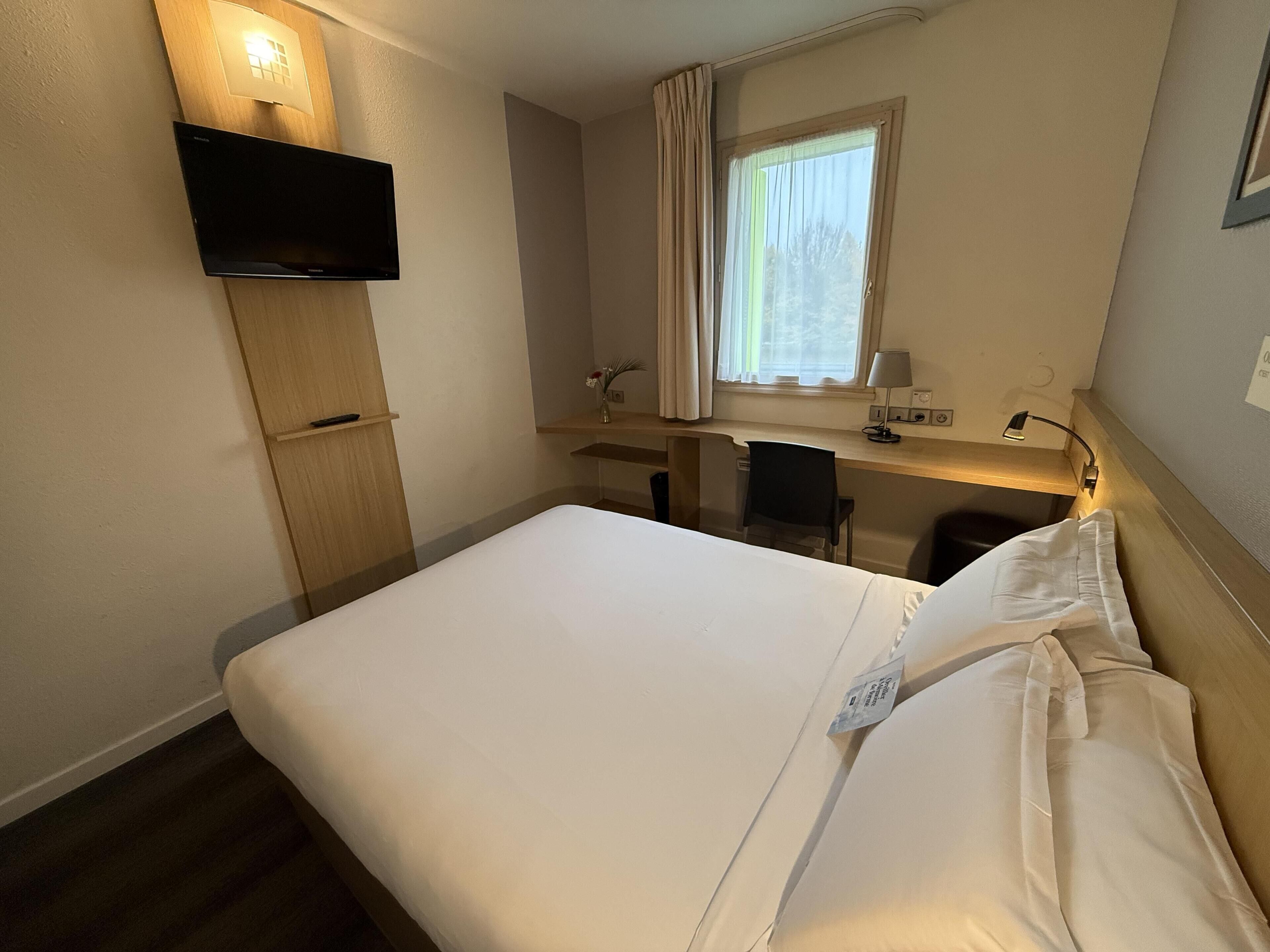 Habitación de hotel con cama, escritorio, silla, televisión y ventana con cortinas.