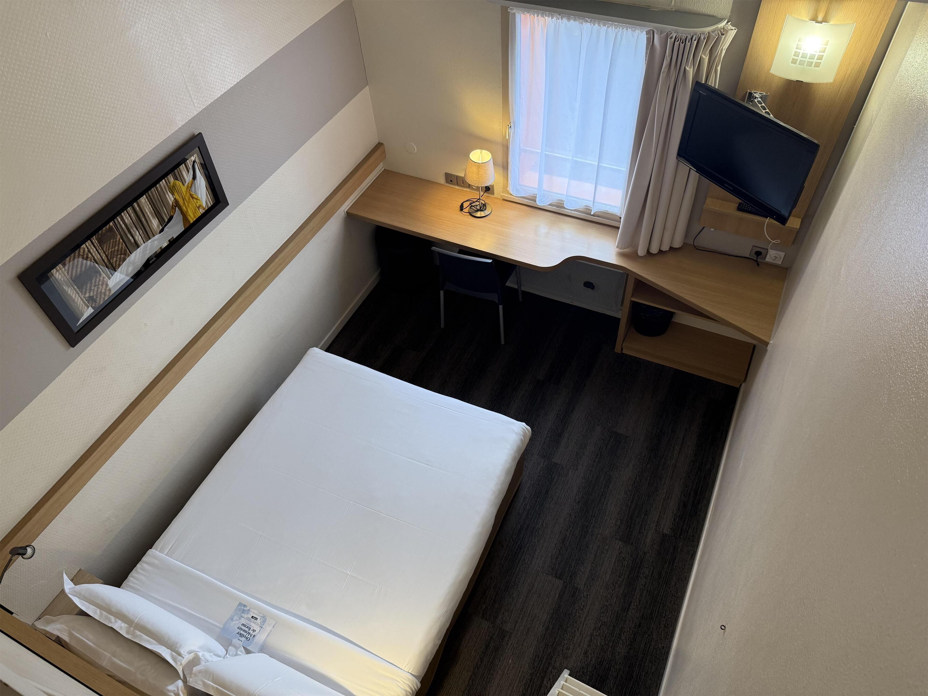A compact room with a bed, a desk, a chair, a small table, a lamp, a TV, and a window with curtains.