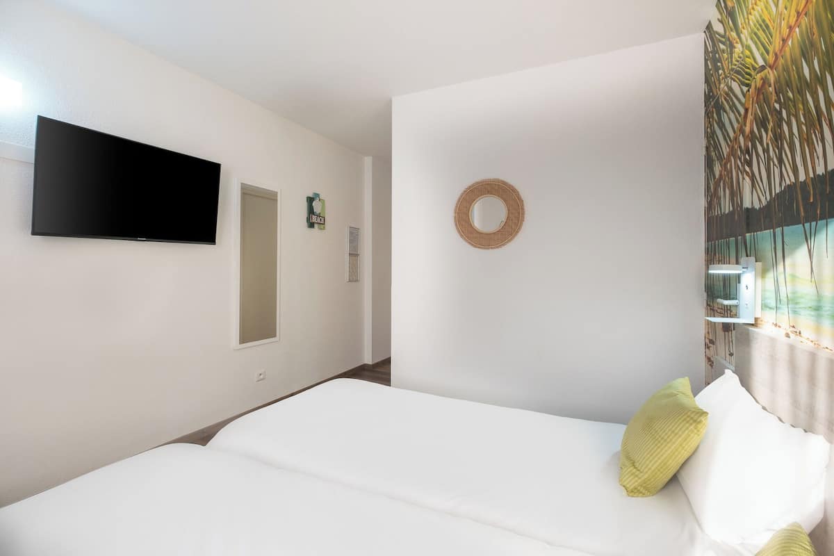 A bedroom with a bed, a flat-screen TV, a door, a wall-mounted mirror, and a window with curtains.