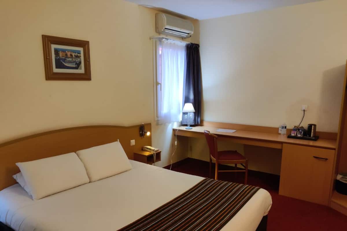 A hotel room with a bed, a desk, a chair, a lamp, and a picture on the wall.