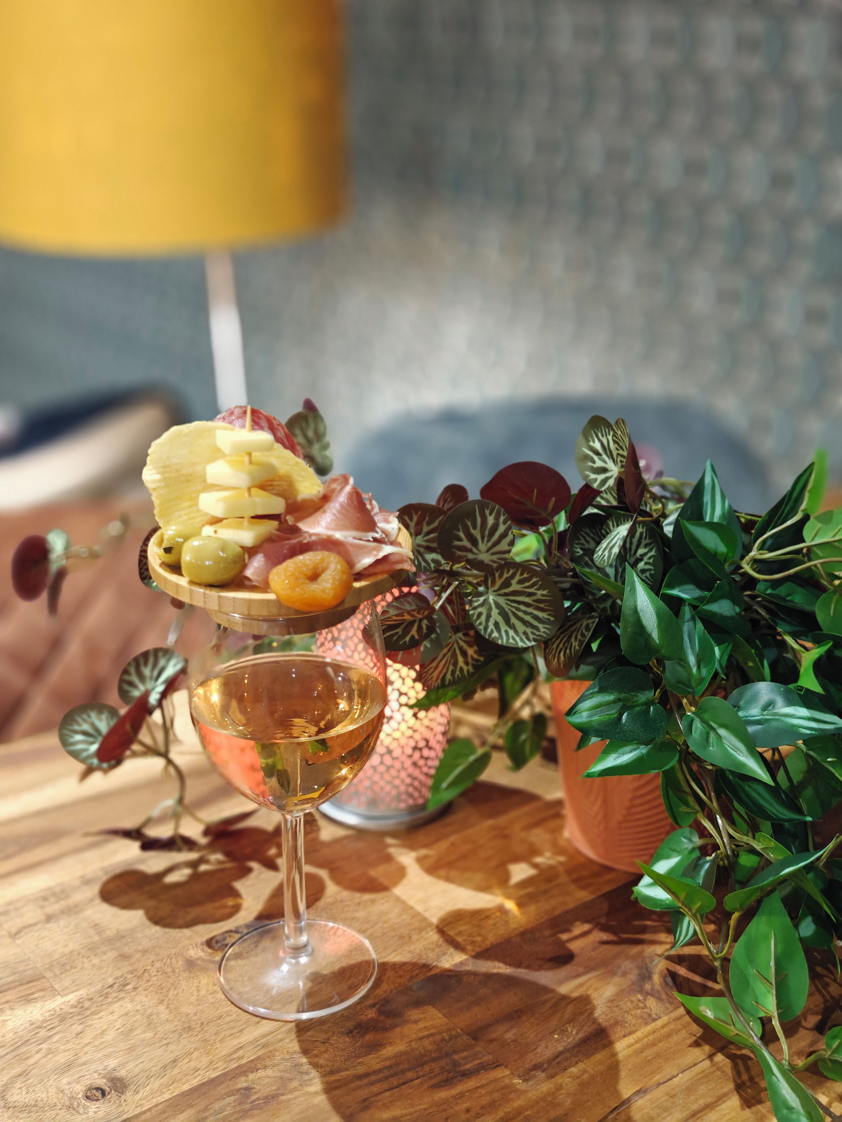 A glass of white wine on a wooden table with a decorative plate of food and a potted plant.