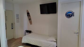 Une petite chambre avec un lit simple, un lit blanc, un téléviseur fixé au mur, une planche de surf et une porte dotée d’une fenêtre ronde.