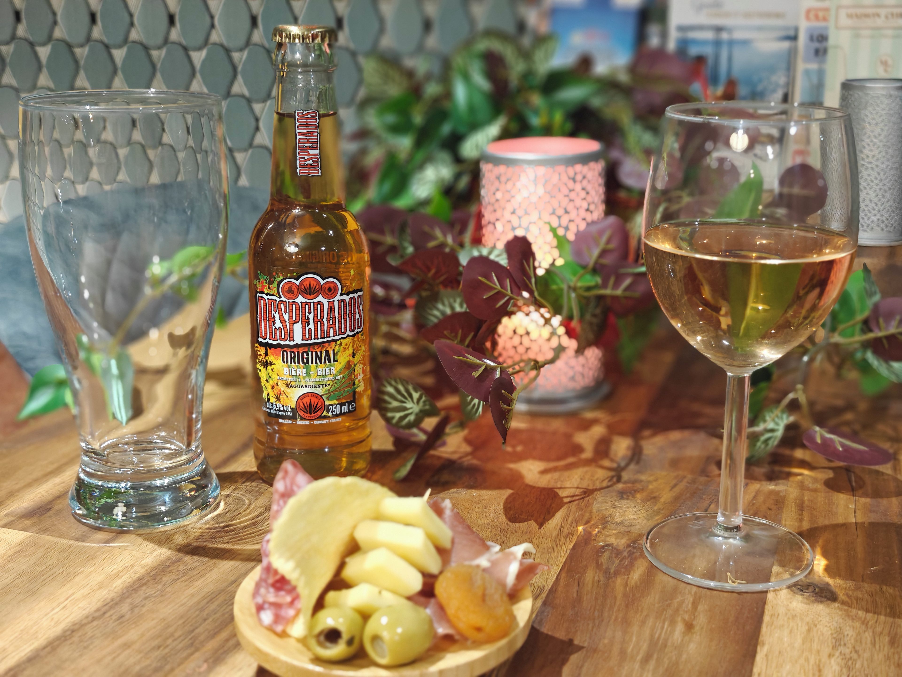 Una botella de cerveza Desperados, una copa de vino, un plato de aceitunas y embutidos sobre una mesa de madera.