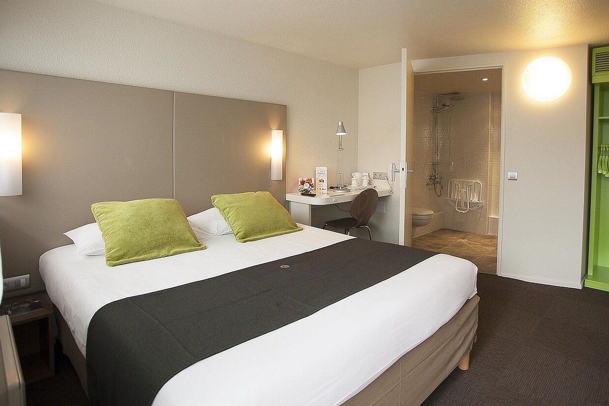 A hotel room with a large bed, a bedside table with a lamp, a green wardrobe, and a shower area.