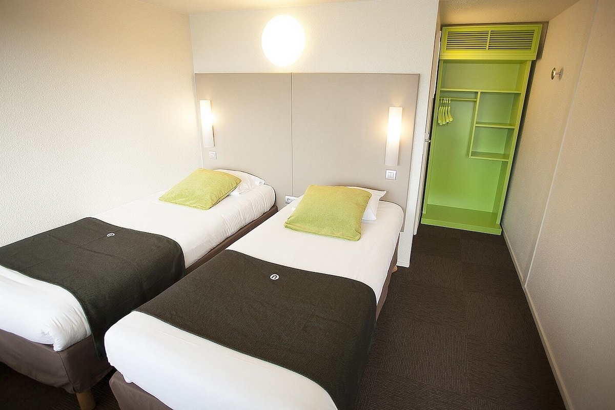 A hotel room with two single beds, each with a pillow and a blanket. There is a green door and a light fixture above the door.