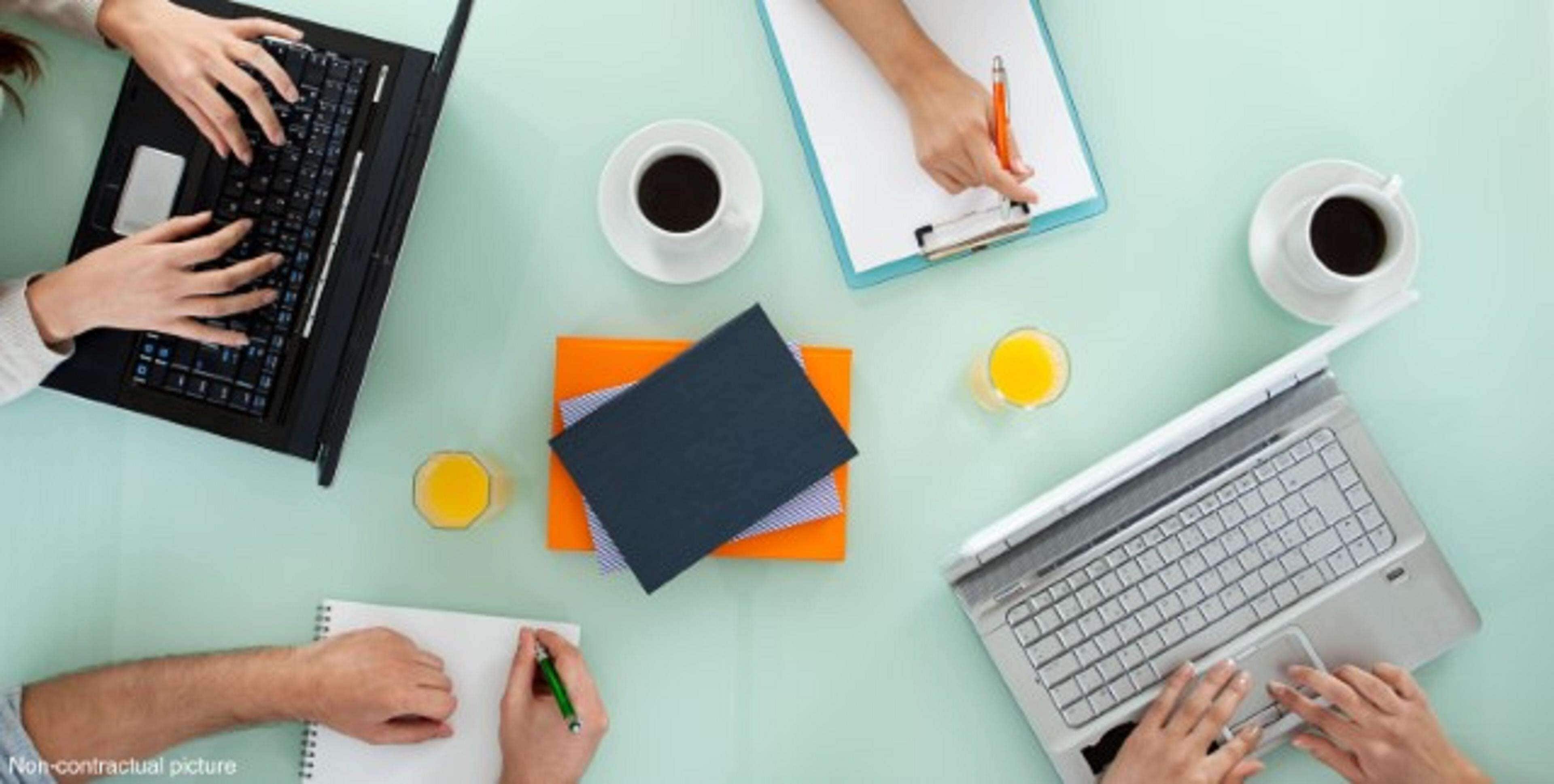 A workspace with two laptops, notebooks, pens, and coffee cups on a light green table.
