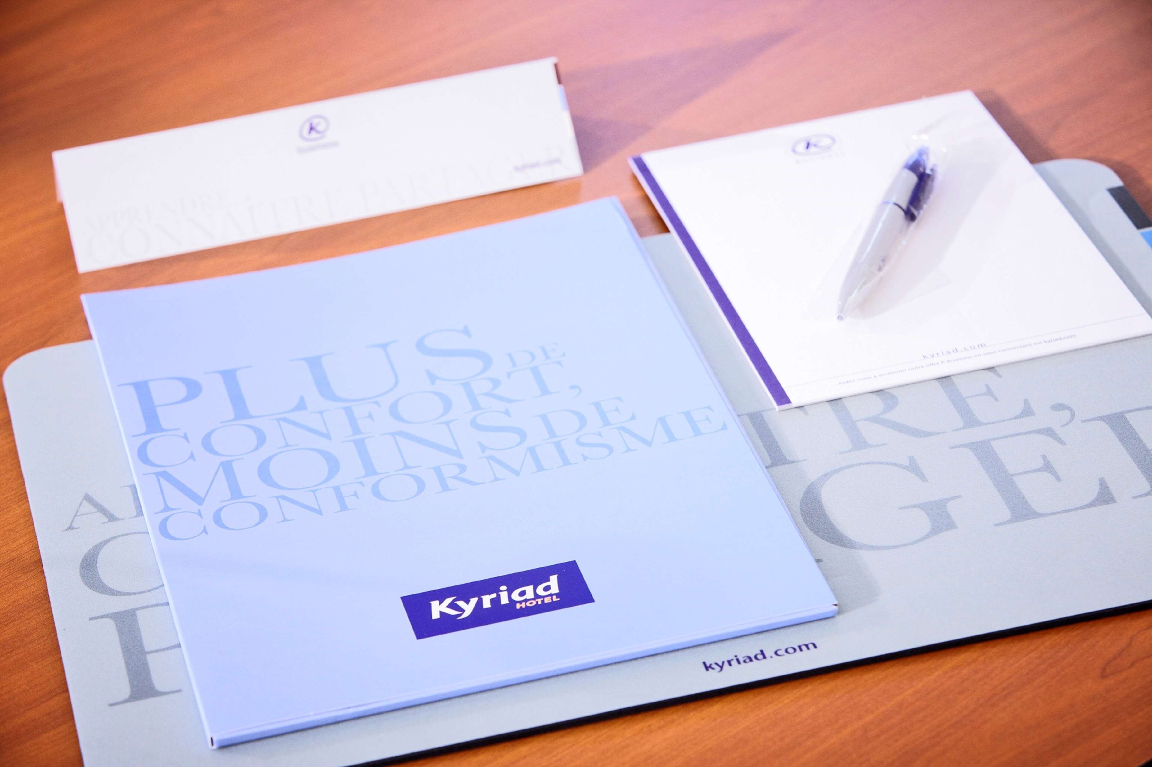 A brochure titled "Plus de Confort, Aimez le Conformisme" from Kyriad Hotel, along with a pen and a business card.