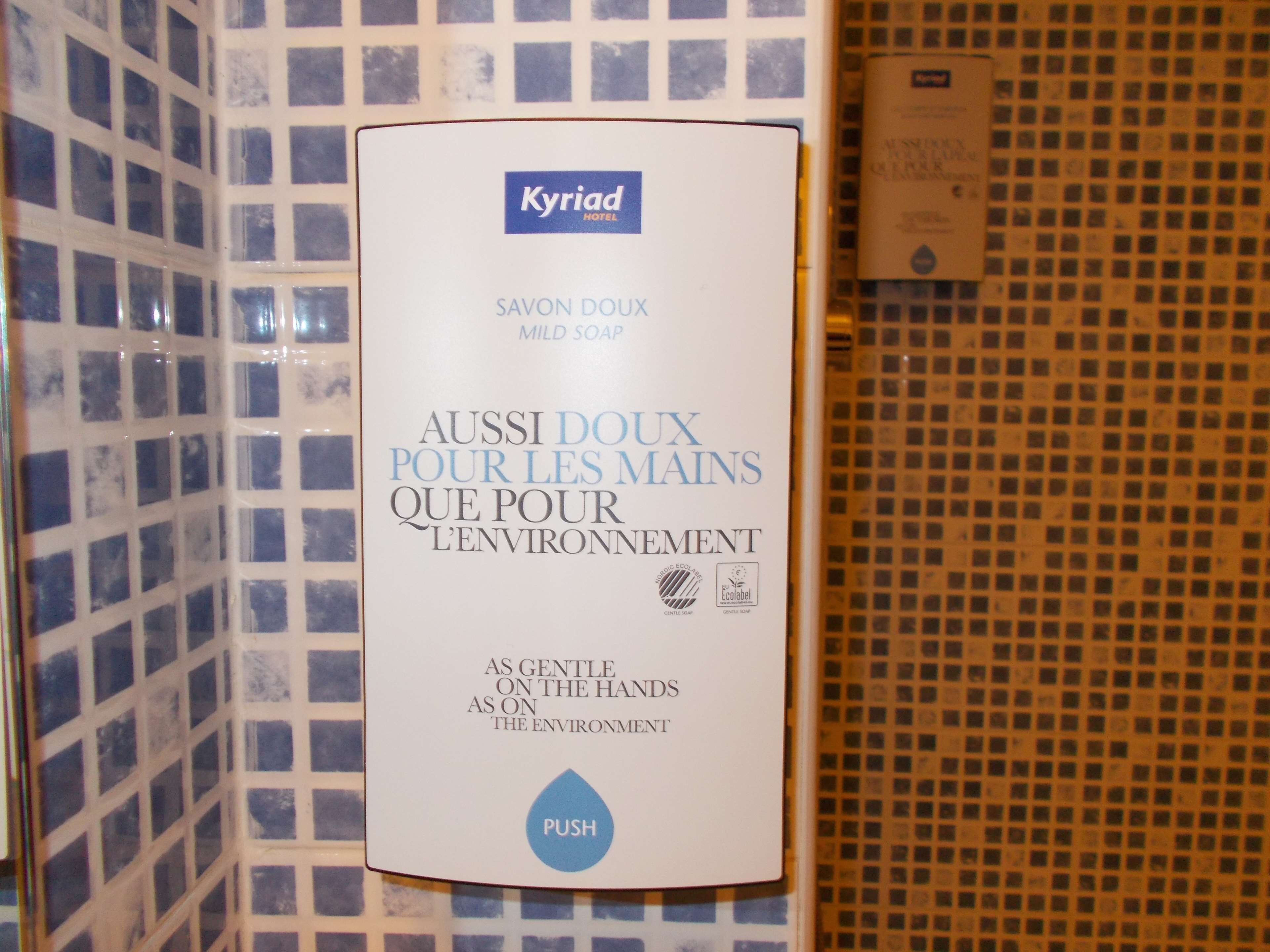 A hotel soap dispenser with the text "Kyriad Hotel" and "Aussi doux pour les mains que pour l'environnement" indicating it's gentle on hands and the environment.