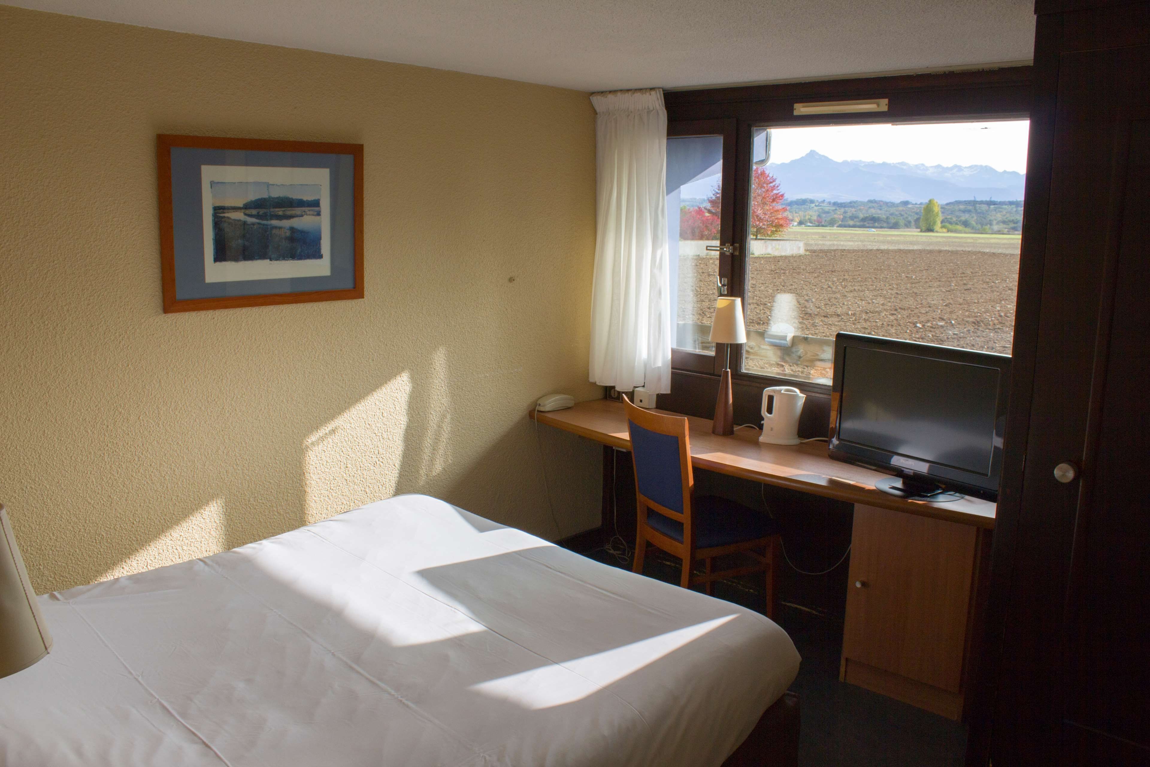 Une chambre d’hôtel avec un lit, un bureau équipé d’un ordinateur, une chaise, une fenêtre donnant sur les montagnes et un tableau encadré au mur.