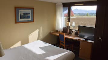 Une chambre d’hôtel avec un lit, un bureau équipé d’un ordinateur, une chaise, une fenêtre donnant sur les montagnes et un tableau encadré au mur.