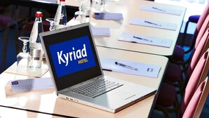 En bærbar computer på et hotelbord viser 'Kyriad Hotel', med et glas vand og en flaske i baggrunden.