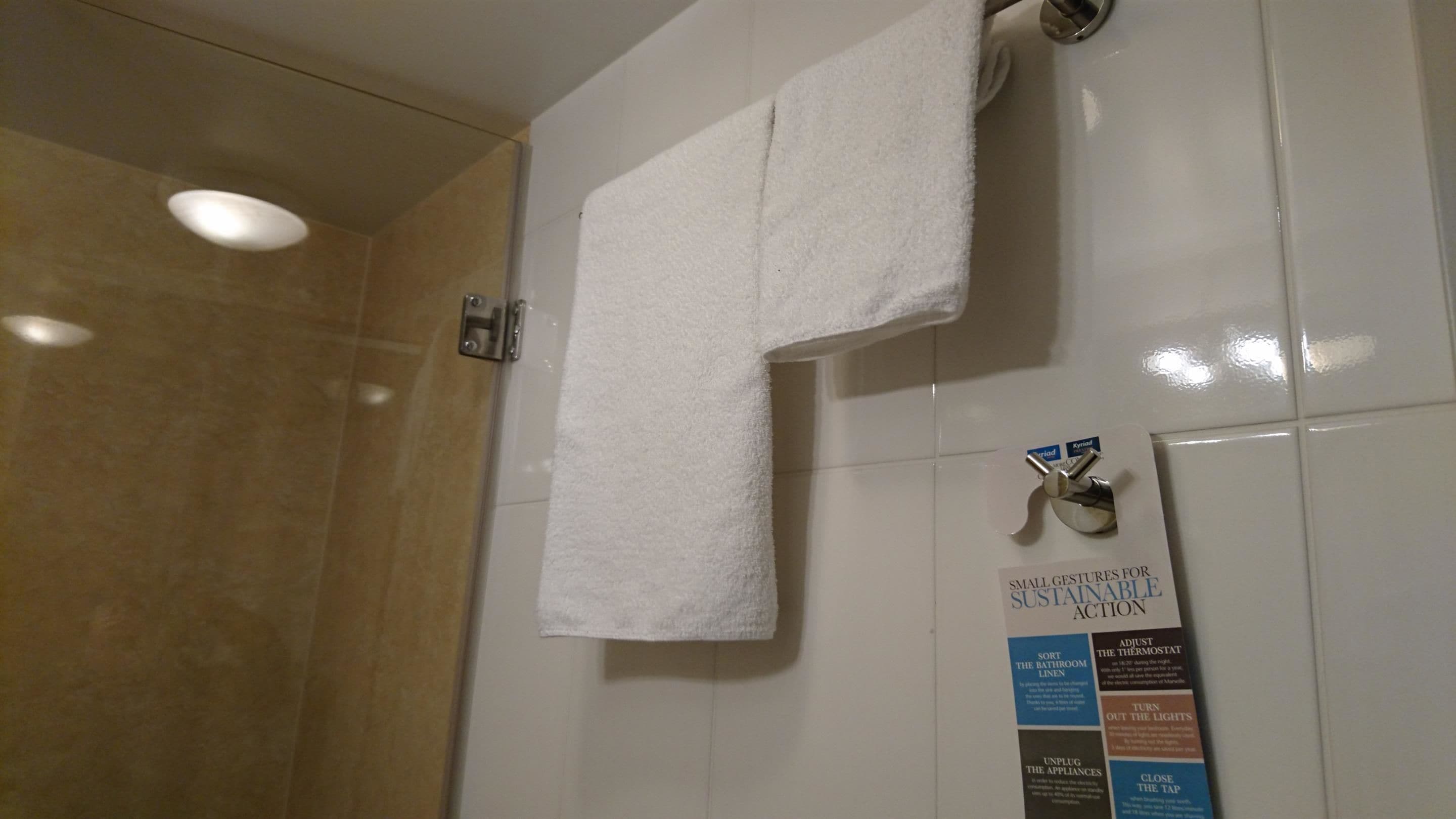 A bathroom with a white towel hanging on a rack, a sign with tips for sustainable action, and a shower area with beige tiles.