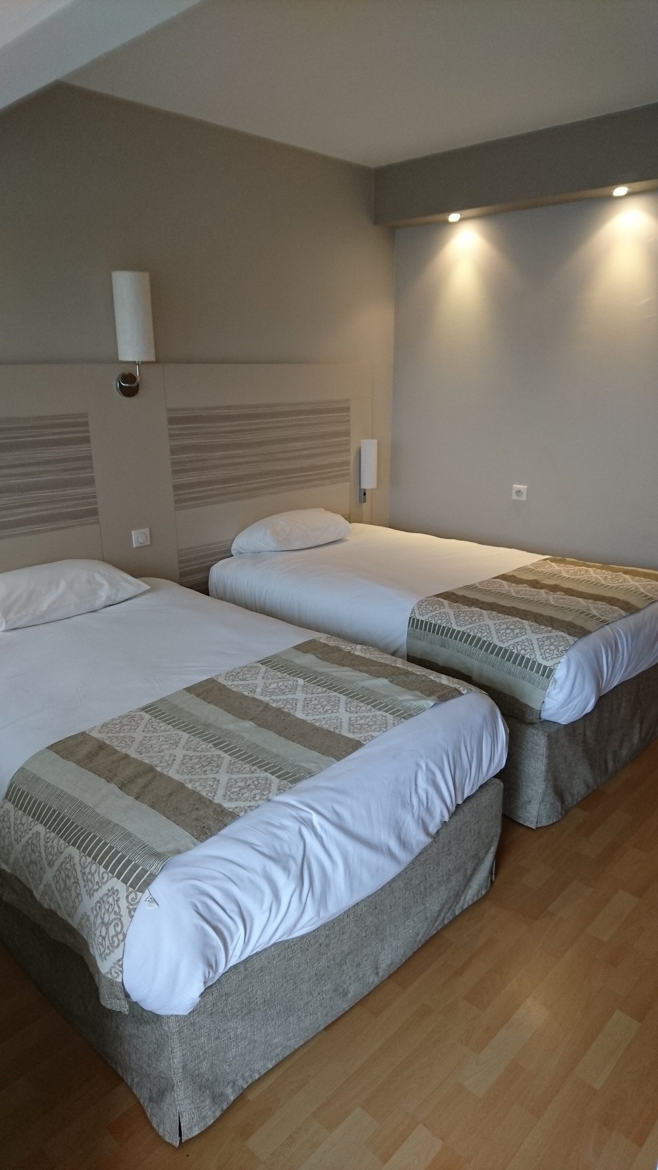 A hotel room with two beds, a wooden floor, and a wall-mounted light fixture.