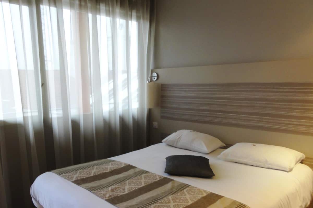 A hotel room with a bed, a bedside lamp, and sheer curtains.