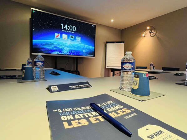 A conference room with a large screen displaying a space-themed wallpaper, a clock, and a table with water bottles, a notebook, and a pen. - Hotel Kyriad Toulouse Sud - Roques (Roques)
