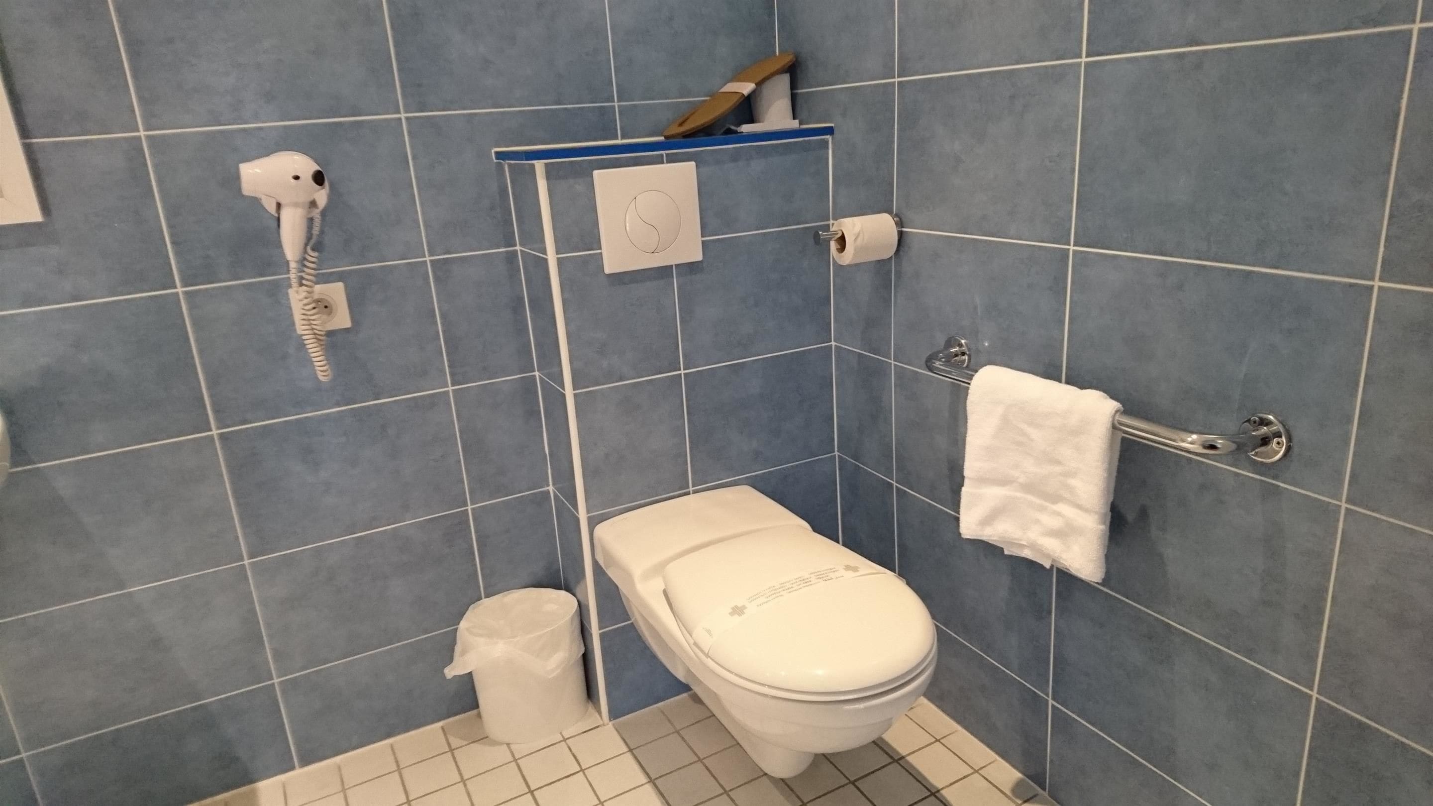 A bathroom with a toilet, a hand dryer, a towel rack, and a trash bin.