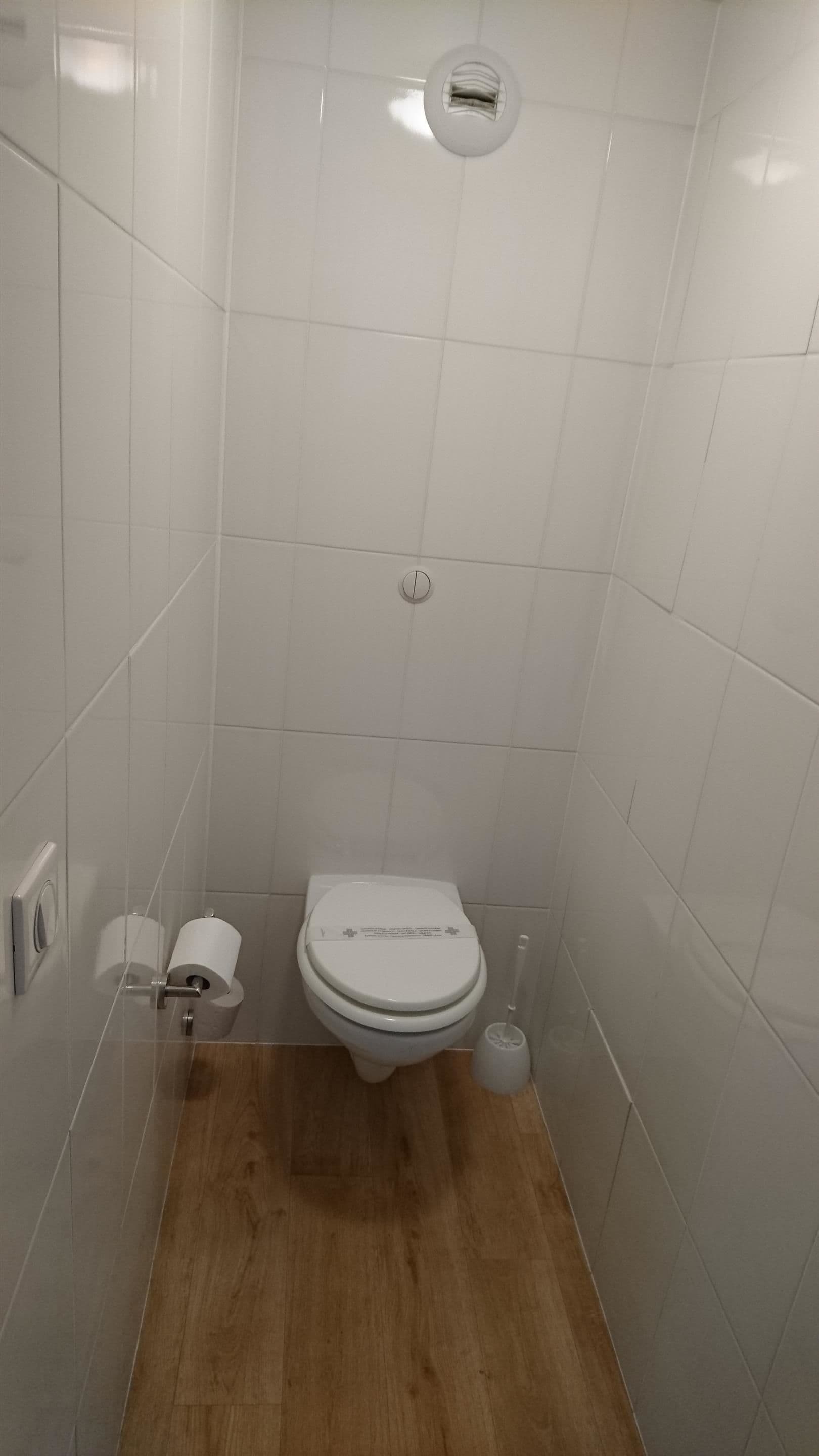 A small, modern bathroom with white tiles, a toilet, a bidet, and a wooden floor.