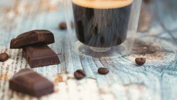 Una taza de café oscuro con espuma, rodeada de granos de café y trozos de chocolate sobre una superficie de madera rústica.