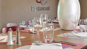 Una mesa de comedor dispuesta con copas de vino, cubiertos y saleros y pimenteros, con una planta en maceta y un letrero que dice "Le Restaurant" al fondo.