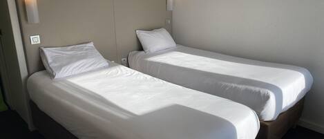 Dos camas individuales con sábanas blancas, un cabecero de color claro y lámparas fijadas a la pared.