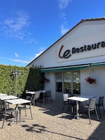 Un área de estar al aire libre con mesas y sillas, un edificio con un letrero que dice 'Le Restaurante' y un seto en el fondo.