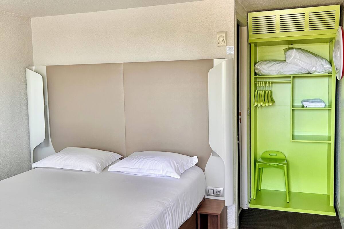 Une chambre d’hôtel avec un lit, une petite table de chevet, une chaise verte et un meuble de rangement vert doté d’un miroir.