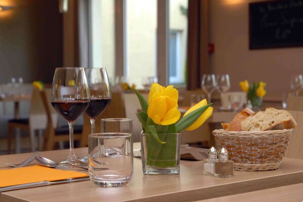 Una mesa de comedor con dos copas de vino, un vaso de agua, un jarrón pequeño con flores amarillas, una cesta de pan y saleros y pimenteros.