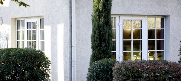Un edificio con dos ventanas, una con marco blanco y otra con una puerta blanca. Un árbol alto y delgado se encuentra frente a la ventana de la puerta.
