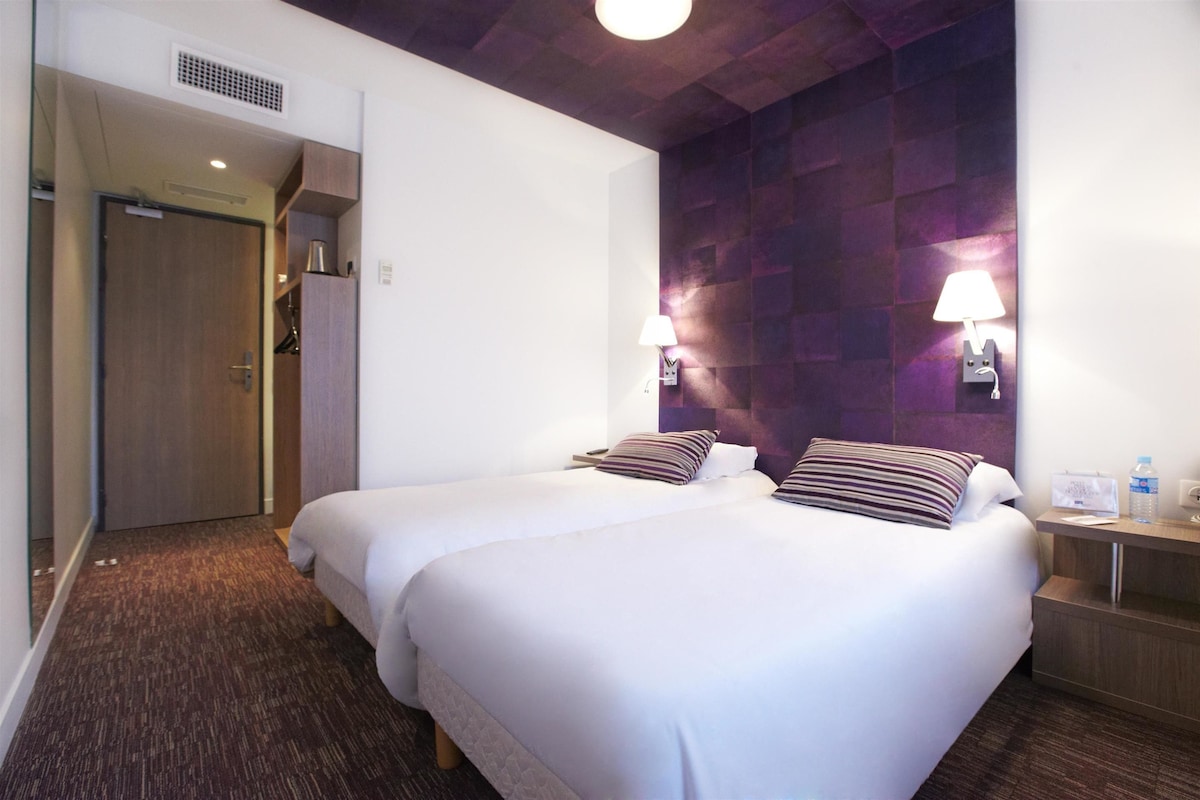 Habitación de hotel con dos camas, cabecero de madera, lámpara de pared y una mesita con una botella y pañuelos.