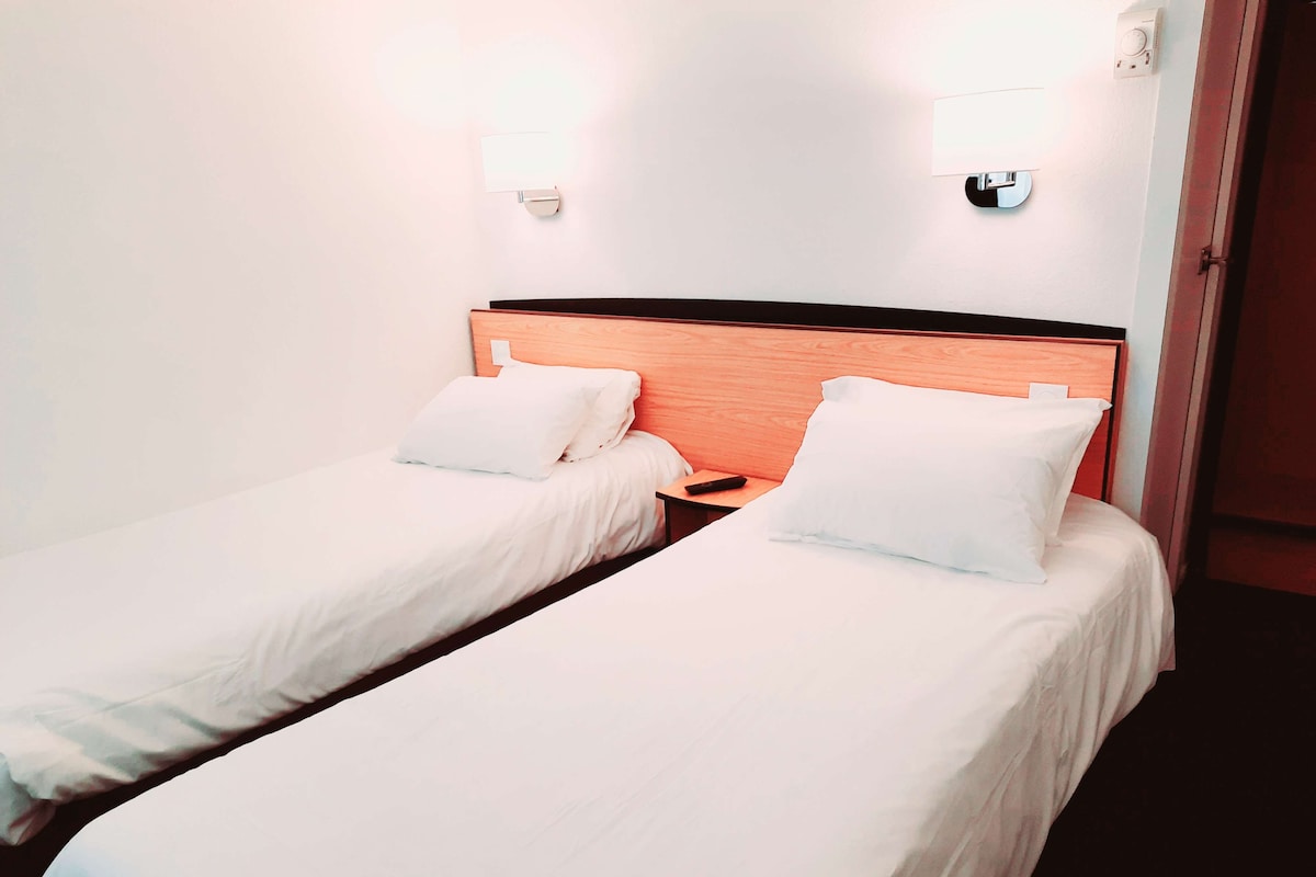 A hotel room with two single beds, each with a wooden headboard, white bedding, and bedside lamps.