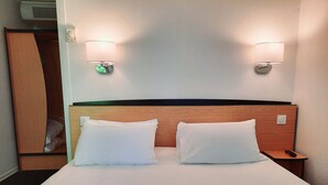 Une chambre d’hôtel avec une tête de lit en bois, deux oreillers blancs, une table de chevet avec une télécommande et des lampes fixées au mur.