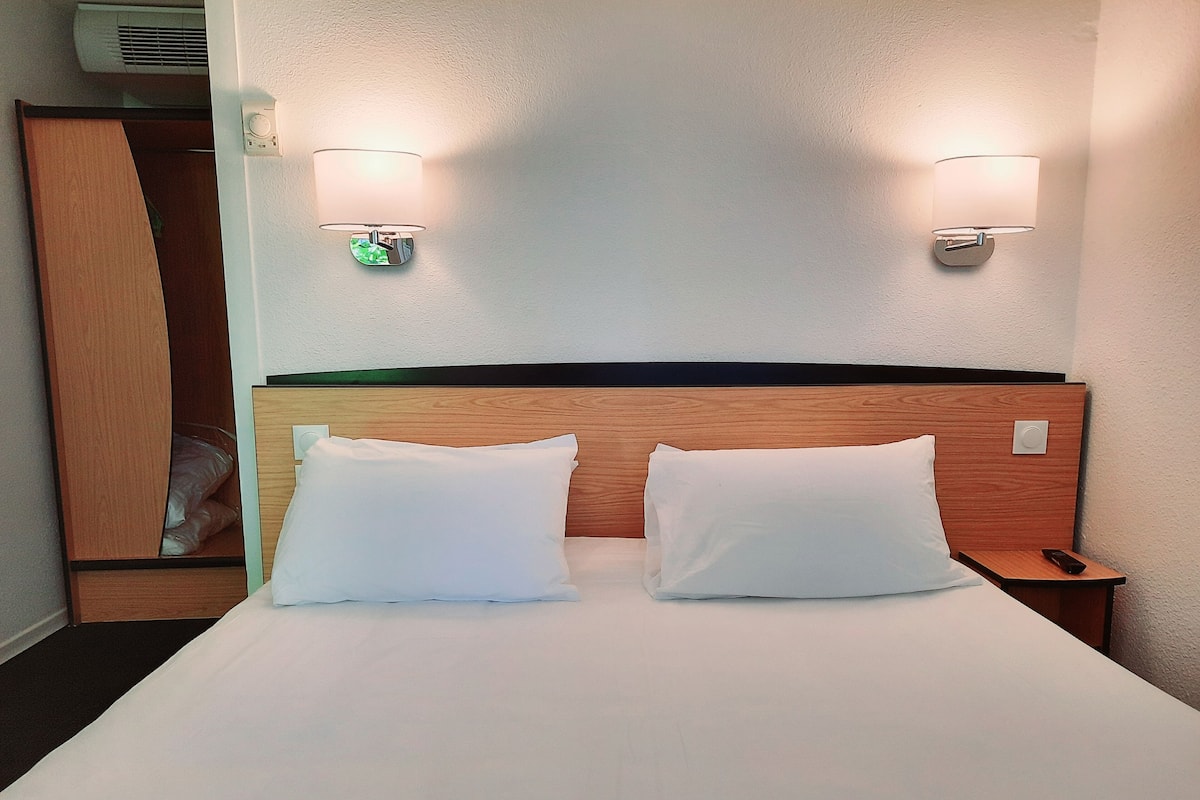 Une chambre d’hôtel avec une tête de lit en bois, deux oreillers blancs, une table de chevet avec une télécommande et des lampes fixées au mur.