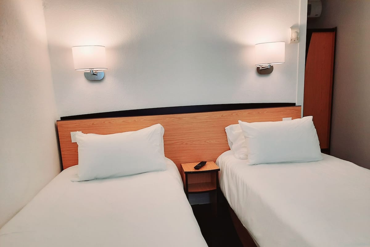 Une chambre d’hôtel avec deux lits simples, chacun doté de draps et d’oreillers blancs. On y trouve deux tables de chevet fixées au mur, avec des lampes, et une tête de lit en bois au-dessus des lits.
