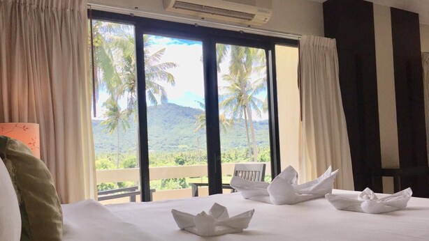 Habitación de hotel con una cama grande, ropa de cama blanca y vistas a palmeras y montañas a través de las ventanas.