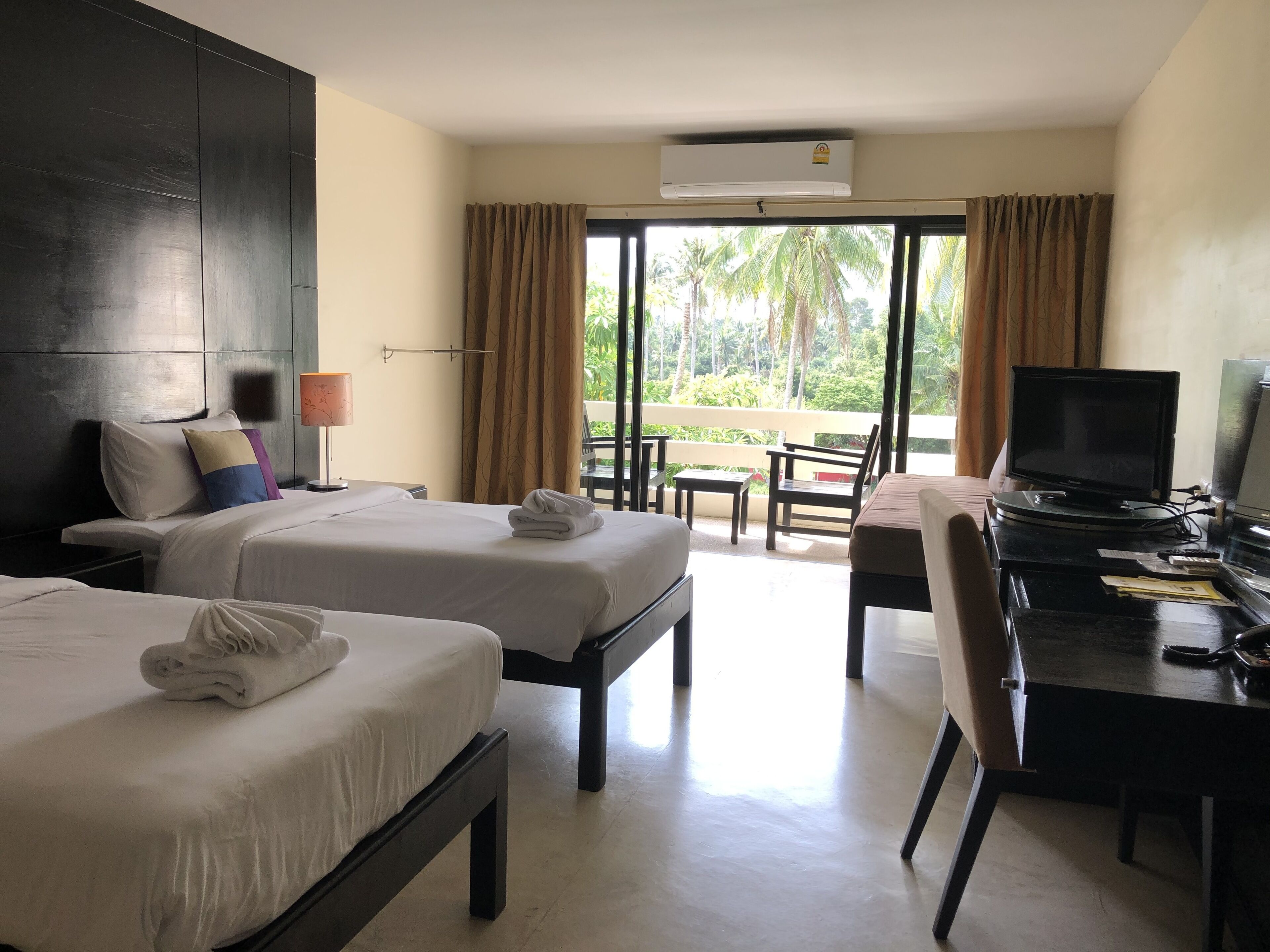 A hotel room with two beds, a desk with a television, and a balcony with a view of palm trees.