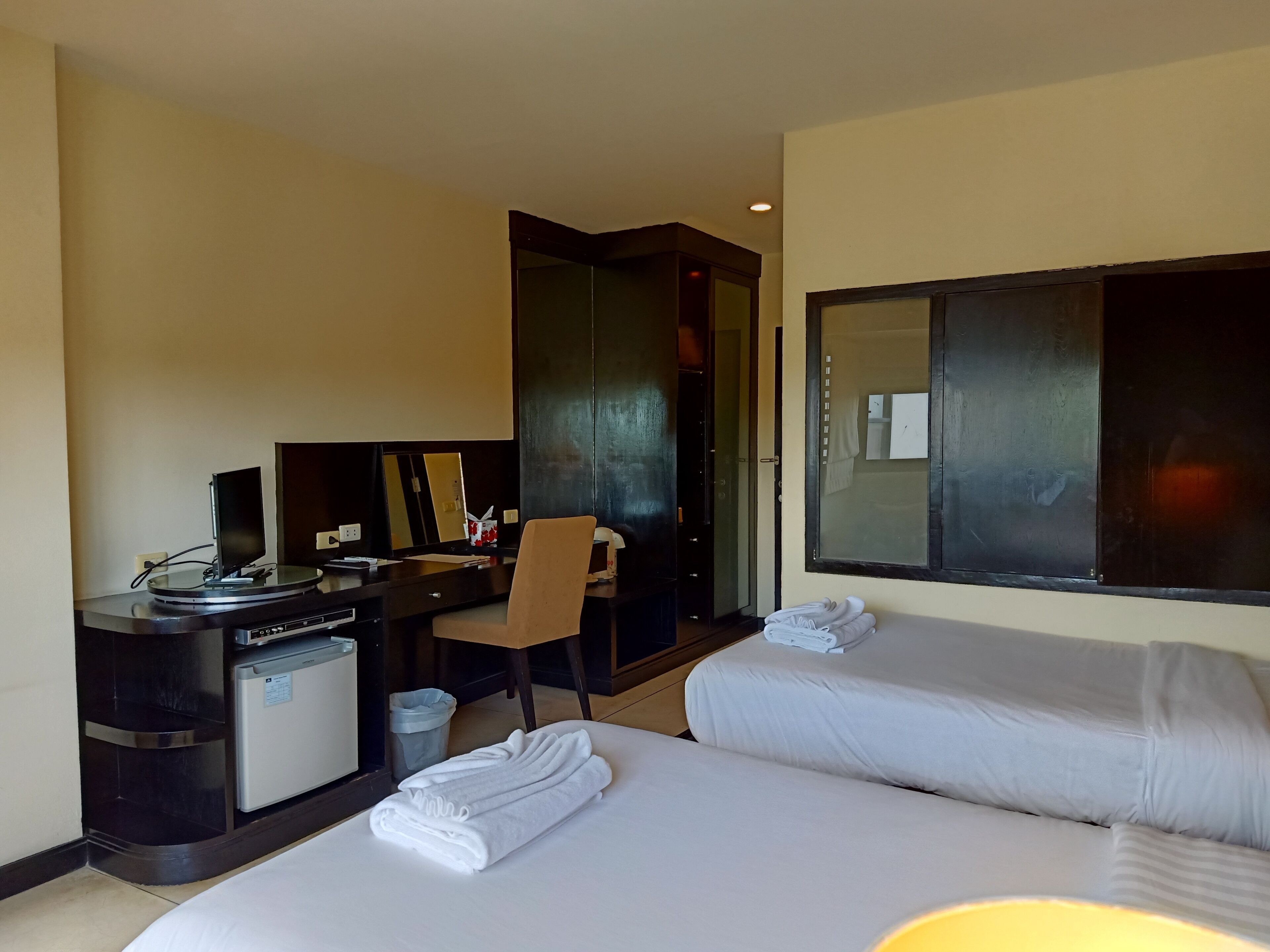 A hotel room with a bed, a desk with a computer, a chair, a small trash bin, and a mirror.