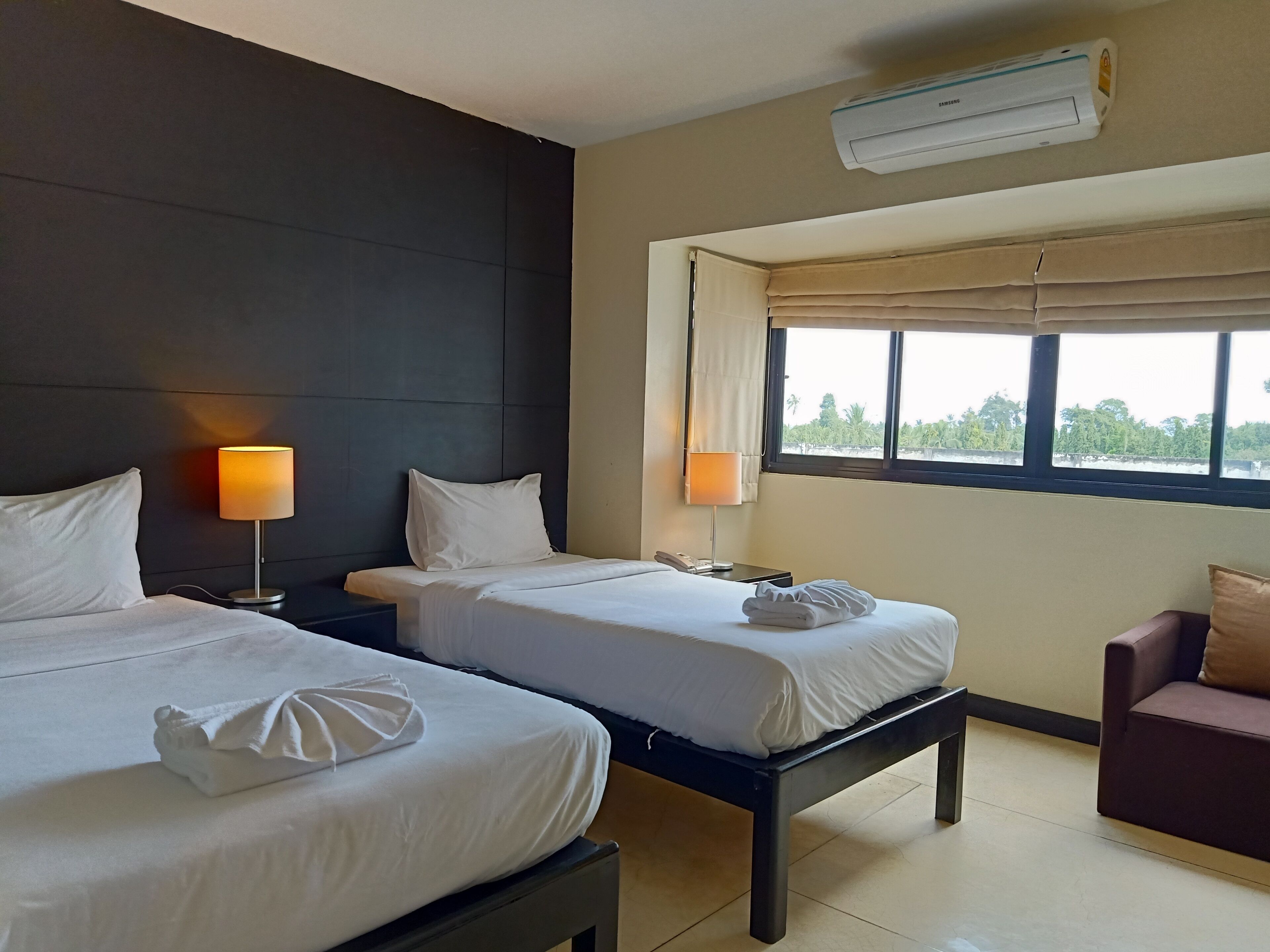 A hotel room with two beds, bedside lamps, a window with a view, and an air conditioning unit.