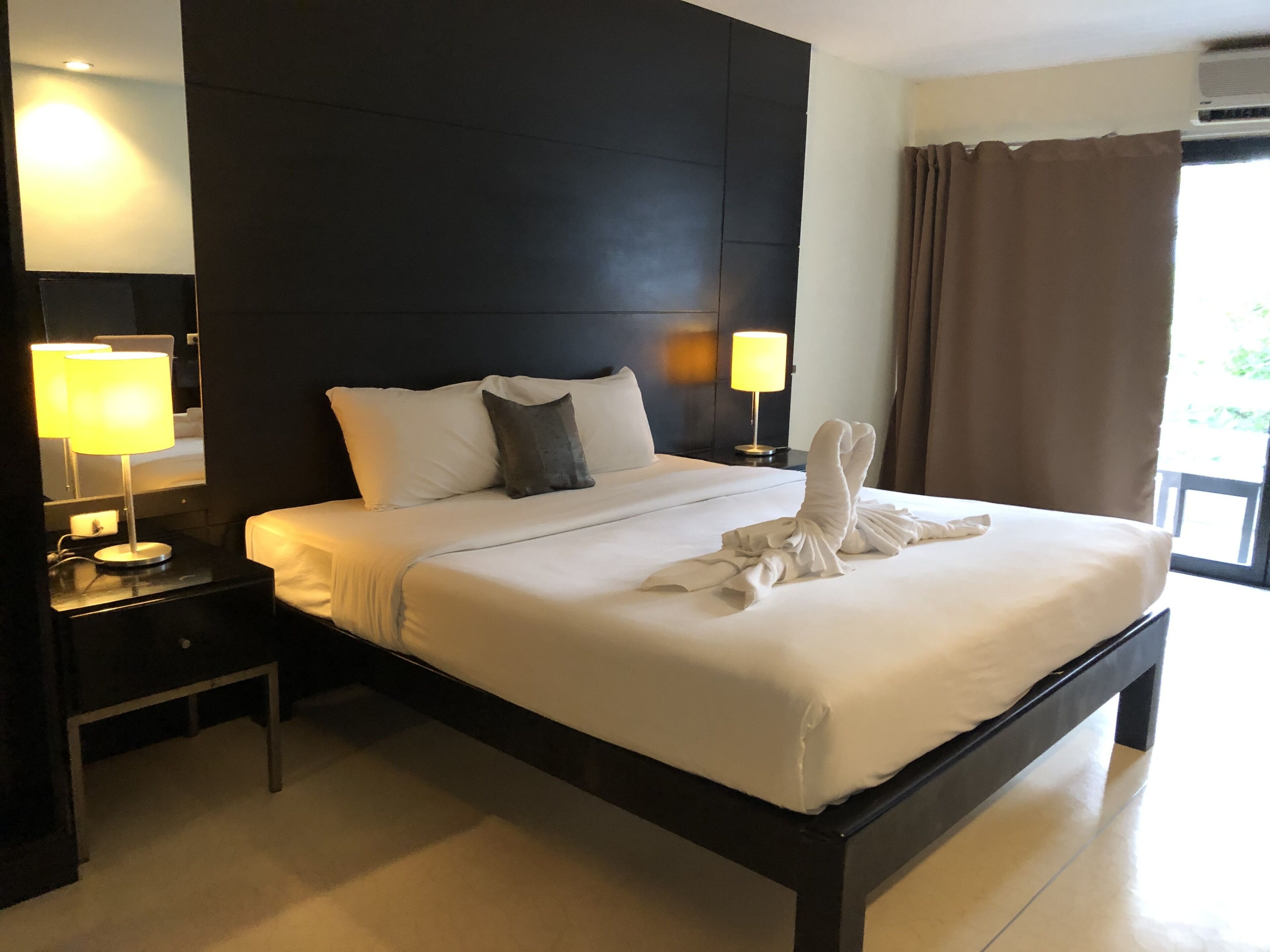A neatly made bed with white linens and a towel arranged in a bow. There are two bedside lamps on black nightstands, a dark headboard, and a window with curtains.