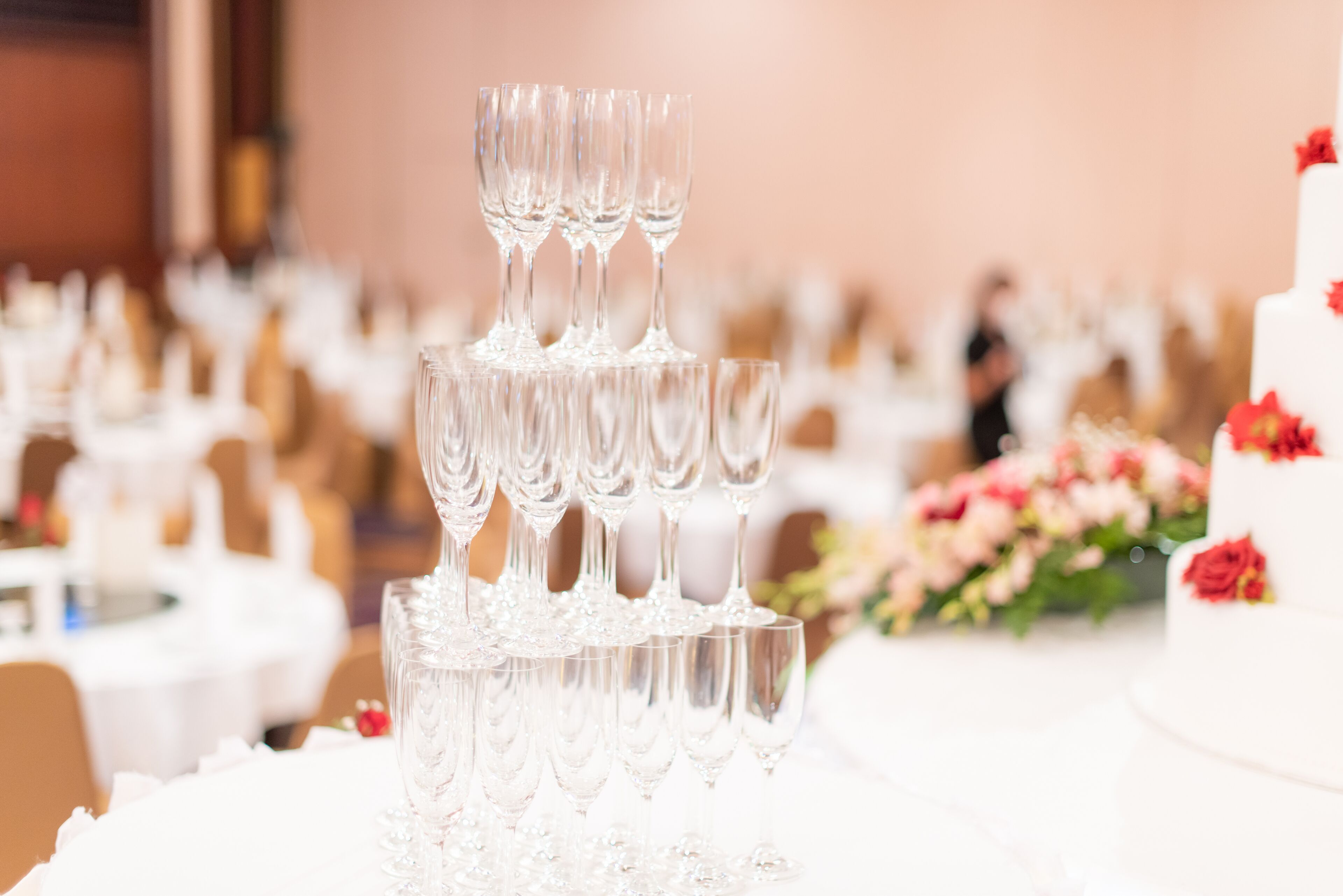 Disposición escalonada de vasos transparentes sobre una mesa, con un pastel blanco y adornos florales al fondo.