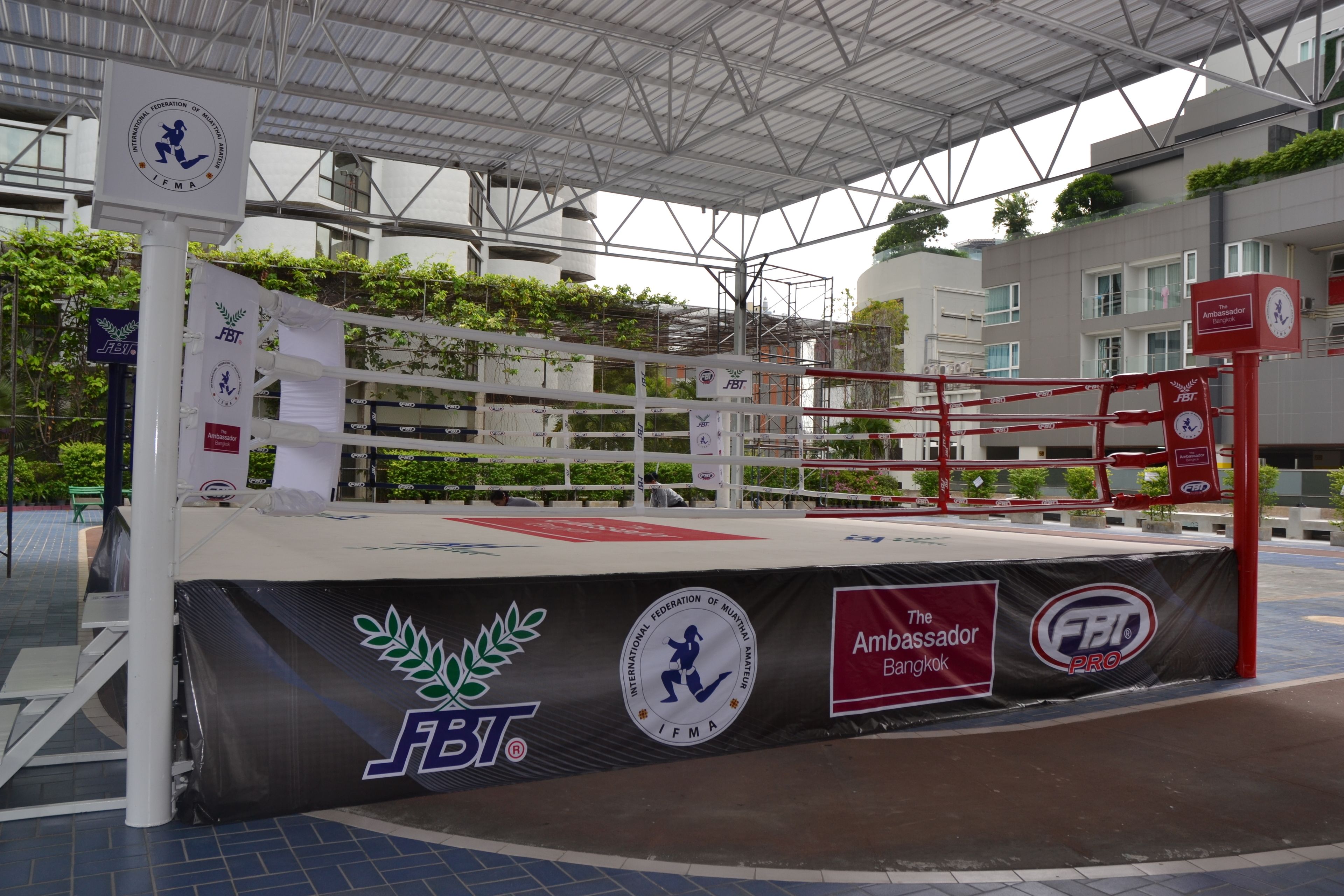 A boxing ring with FBT and IFMA logos, surrounded by a covered area with a metal roof.
