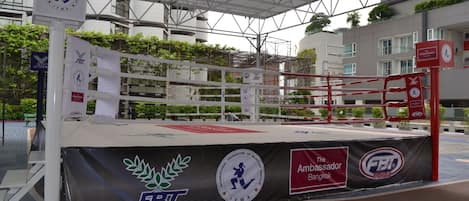 Um ringue de boxe com os logotipos FBT e IFMA, rodeado por uma área coberta com telhado de metal.
