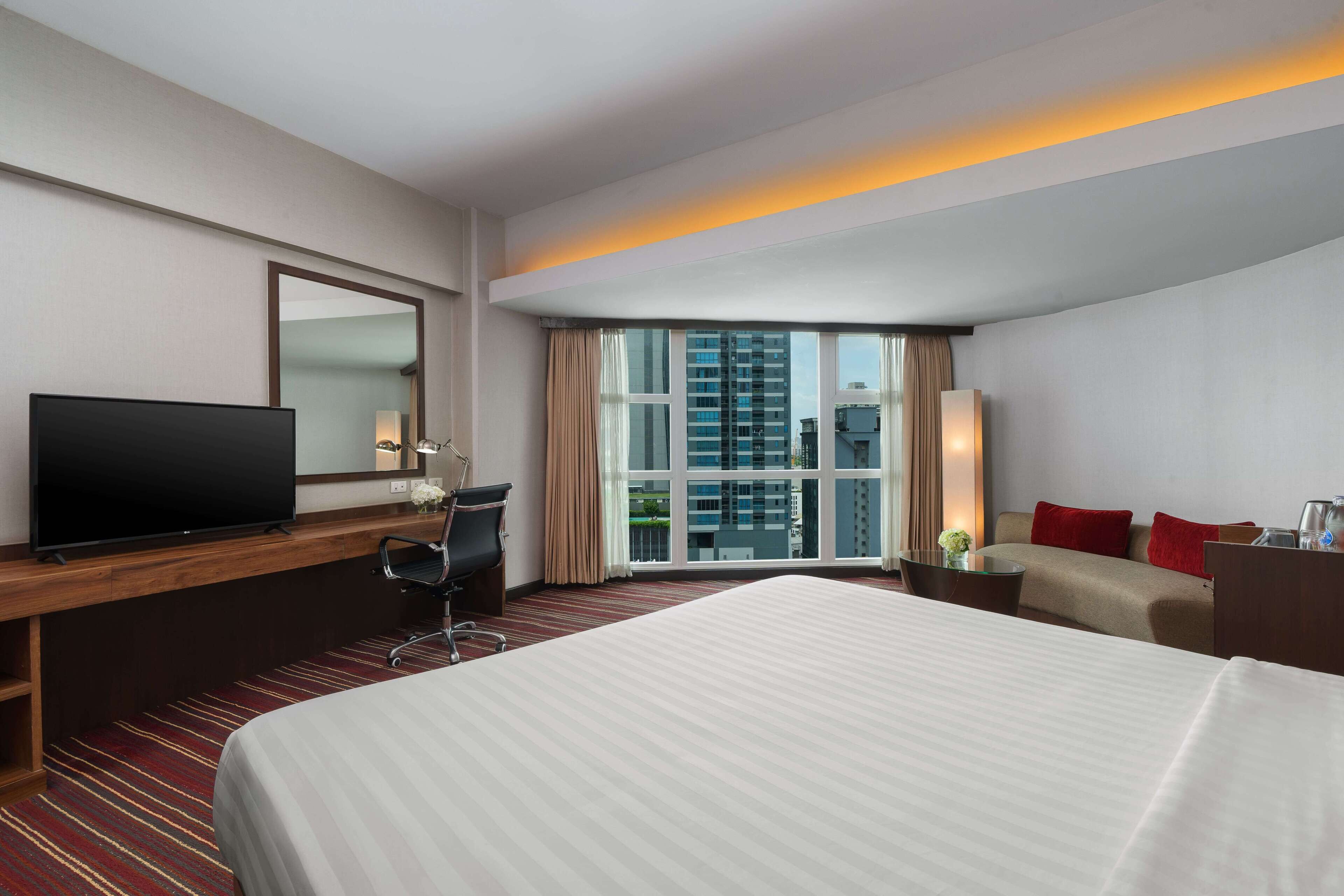 Habitación de hotel con una cama grande, un escritorio con televisor de pantalla plana, una silla, un sofá y una ventana con vista a la ciudad.