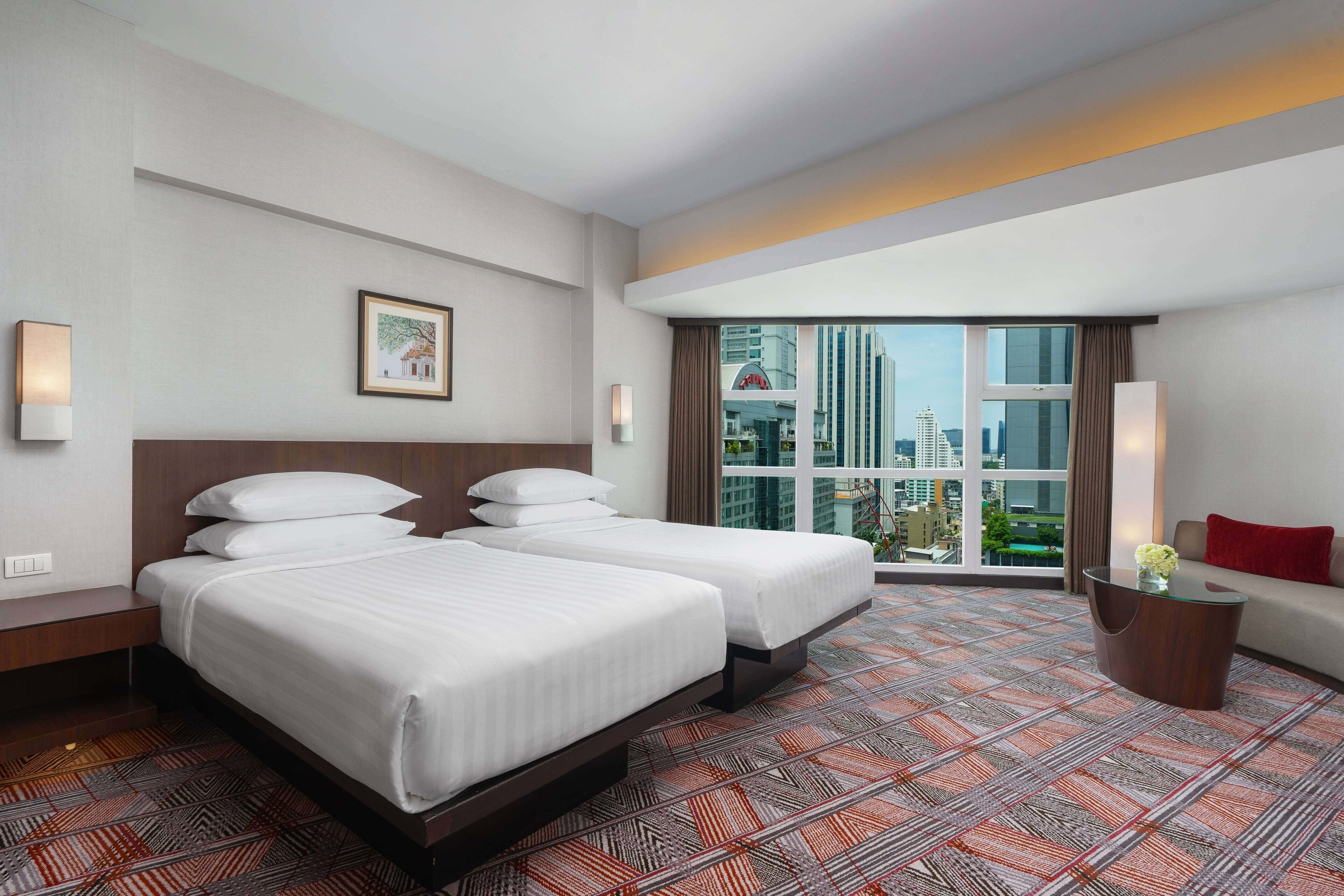 A hotel room with two beds, a large window with a city view, a small table with a vase, and a red pillow on a sofa.