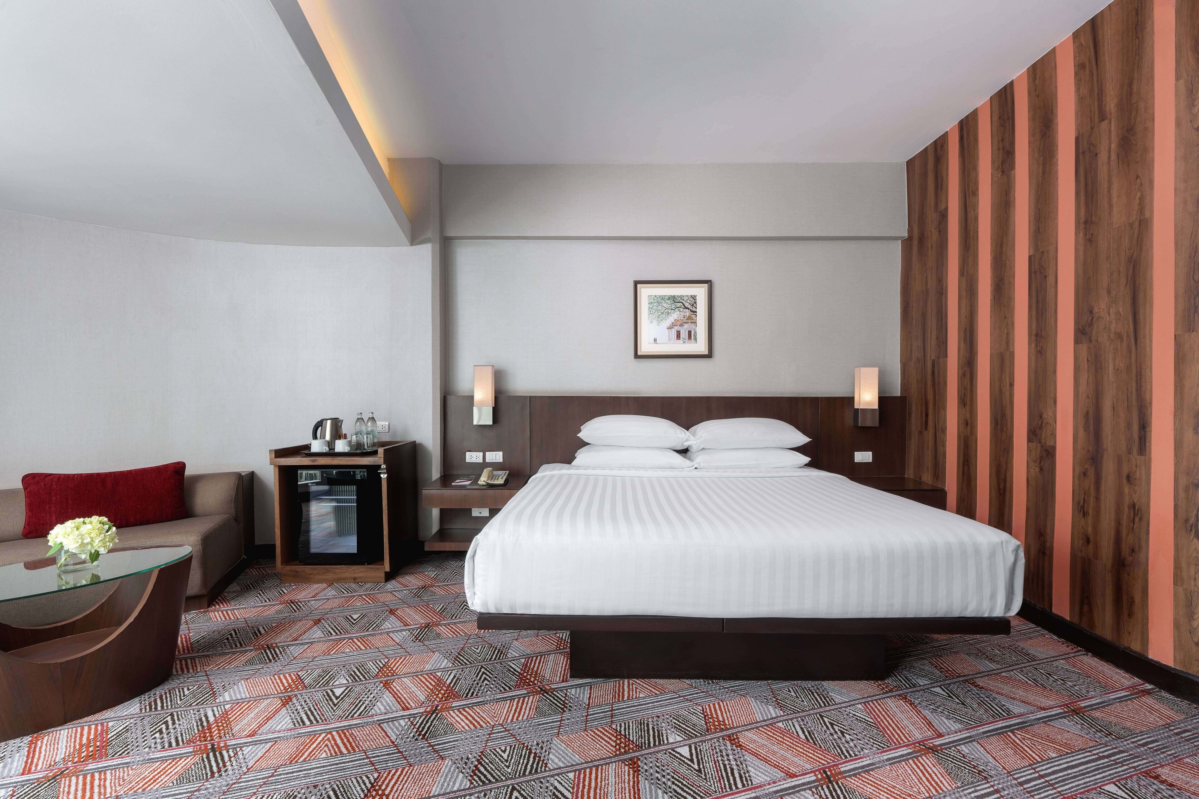 A hotel room with a bed, bedside tables, a desk, a chair, a sofa, and a patterned carpet.