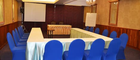 Un salón de conferencias con una larga mesa, sillas azules, una pantalla de proyector y una pizarra.